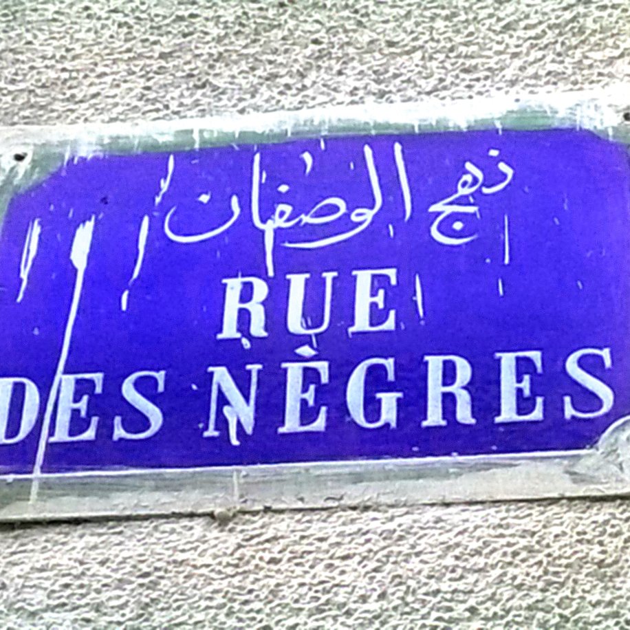 The image shows a street sign. The sign is rectangular and features a blue background with white lettering. It includes text in French that reads "RUE DES NÈGRES," along with Arabic script above it. The sign appears to be weathered, with some signs of wear and tear.