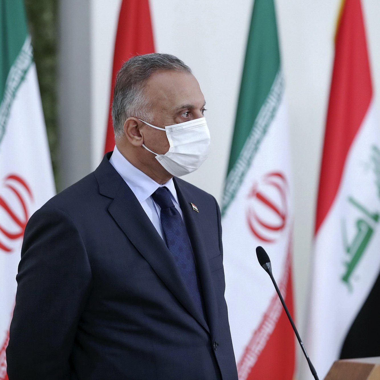 L'image montre un homme en costume, portant un masque facial. Il se tient devant plusieurs drapeaux, dont ceux de l'Iran et de l'Irak. Son expression semble sérieuse, et il semble être en train de participer à une conférence ou une réunion. Le décor est formel, suggérant un cadre politique ou diplomatique.