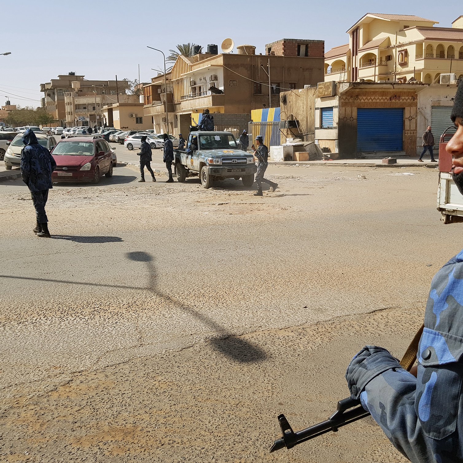 L'image montre une scène urbaine où des policiers armés sont présents dans une rue. On peut voir plusieurs membres des forces de l'ordre, certains portant des uniformes camouflés et des masques, tout en étant attentifs à leur environnement. Il y a des véhicules de police, ainsi que des voitures stationnées à proximité. En arrière-plan, on aperçoit des bâtiments de style architectural varié. L'atmosphère semble tendue, évoquant un contexte de sécurité renforcée.
