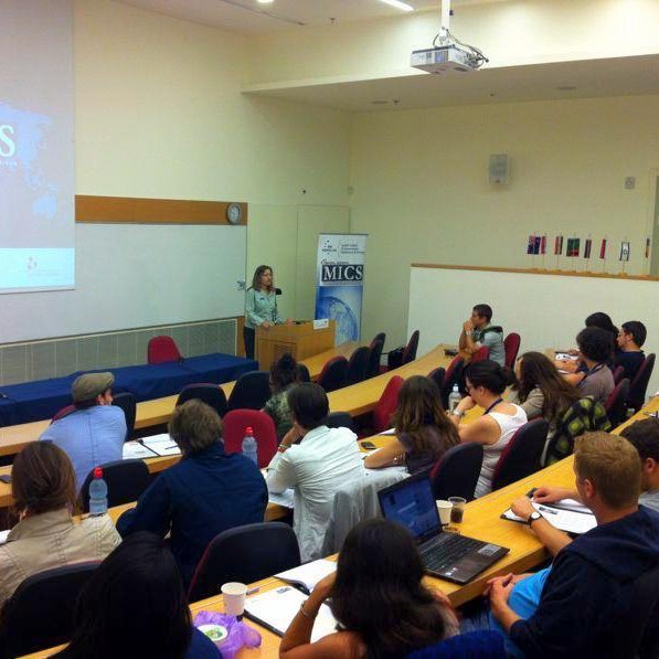 L'image montre une salle de classe ou un auditorium où une présentation est en cours. À l'avant, une intervenante se tient près d'un pupitre, probablement en train de s'adresser à l'auditoire. En arrière-plan, un écran affiche le mot "MICS" accompagné d'une carte du monde. Le public, composé d'une douzaine de personnes, semble attentif et prend des notes sur des ordinateurs portables ou des bloc-notes. La disposition des sièges est en rangées, et quelques personnes sont assises avec des tasses et des boissons. L'ambiance semble académique et interactive.