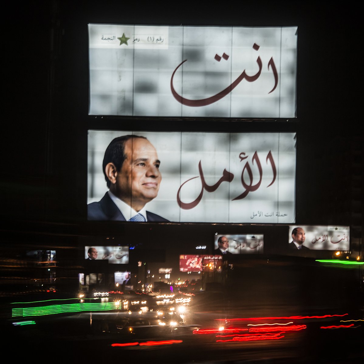 L'image montre un grand panneau d'affichage lumineux à la nuit tombante. Sur ce panneau, on peut voir un portrait d'un homme en costume. Les mots arabes "أنت الأمل" et "أنت" sont écrits en grandes lettres, indiquant un message fort. En bas, il y a une autre image de l'homme. L'environnement est animé, avec des lumières de voitures qui passent en bas, créant un effet de mouvement et une ambiance urbaine nocturne.