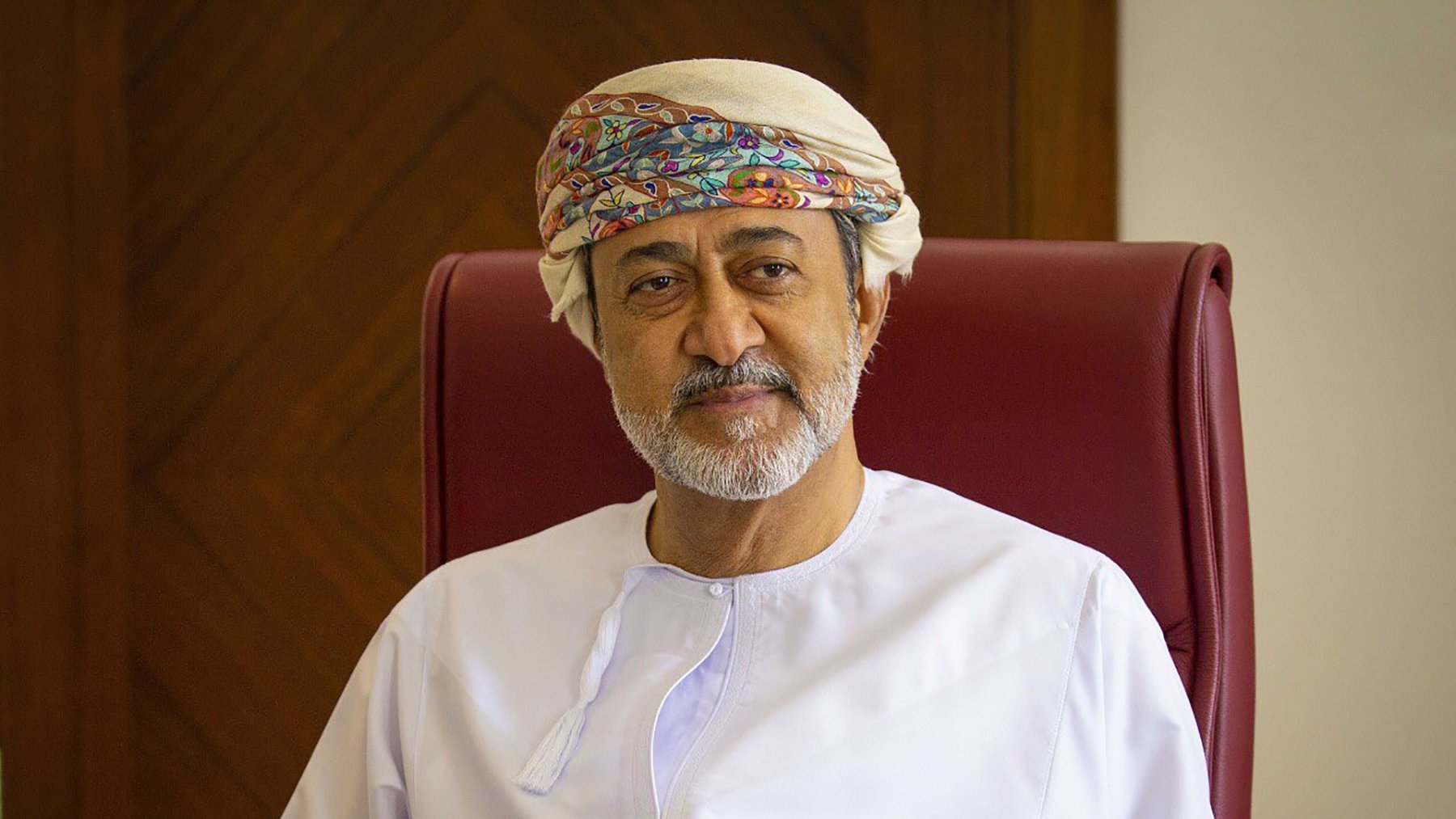 الصورة تظهر شخصًا يجلس على كرسي. يرتدي الملابس التقليدية العمانية، حيث يظهر بجلابية بيضاء وعصابة رأس مزخرفة. في يده يوجد خنجر عماني تقليدي. خلفه خلفية خشبية بسيطة. يعكس تعبير الوجه الهدوء والثقة.