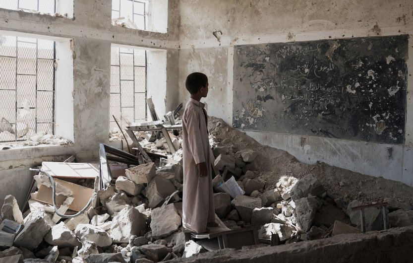 در این تصویر، یک پسر جوان در یک کلاس درس ویران شده ایستاده است. دیوارها و سقف کلاس آسیب دید‌ه‌اند و بخش‌هایی از زیرساخت‌ها به طور کامل تخریب شده‌اند. در اطراف او، زباله‌ها و آوارهای ساختمانی وجود دارد. یک تابلو سیاه با نوشته‌هایی بر روی دیوار وجود دارد، اما وضعیت آن نیز نامناسب است. نور از پنجره‌های شکسته به فضای داخلی می‌تابد و حس ناامیدی و ویرانی را منتقل می‌کند.