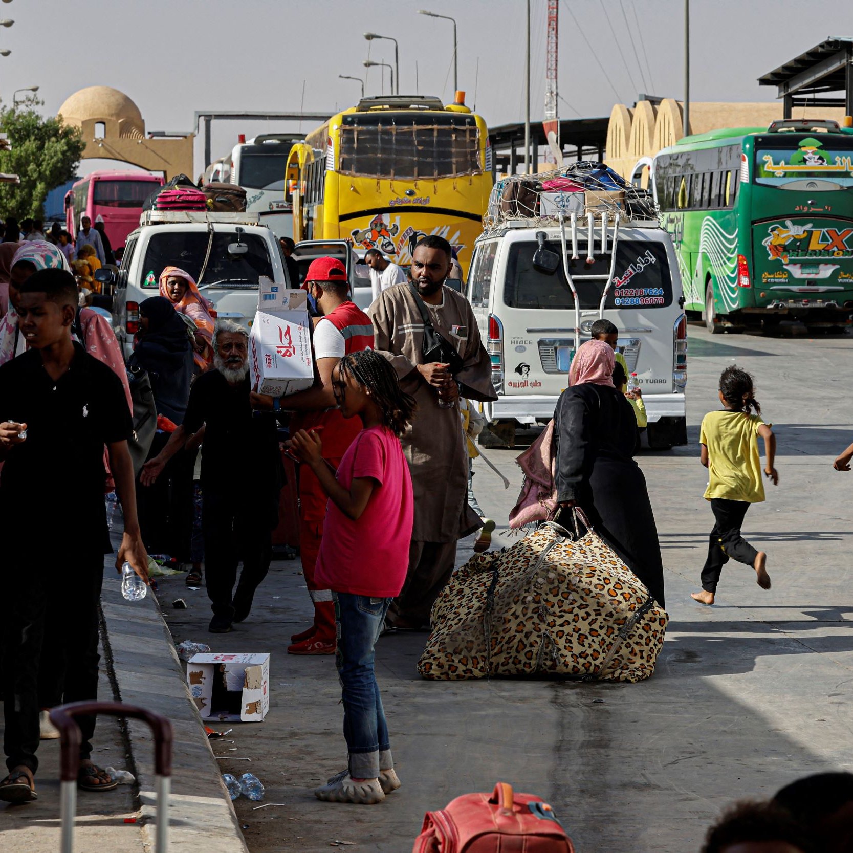 L'image montre une scène animée dans une gare routière ou un terminal. On peut voir plusieurs bus stationnés, avec des passagers qui se déplacent dans différentes directions. Certaines personnes portent des bagages, tandis que d'autres semblent attendre ou se préparer à monter dans un bus. L'atmosphère est dynamique, avec une variété de personnes, notamment des familles et des enfants. Le décor extérieur montre un espace public ensoleillé avec des bâtiments en arrière-plan.