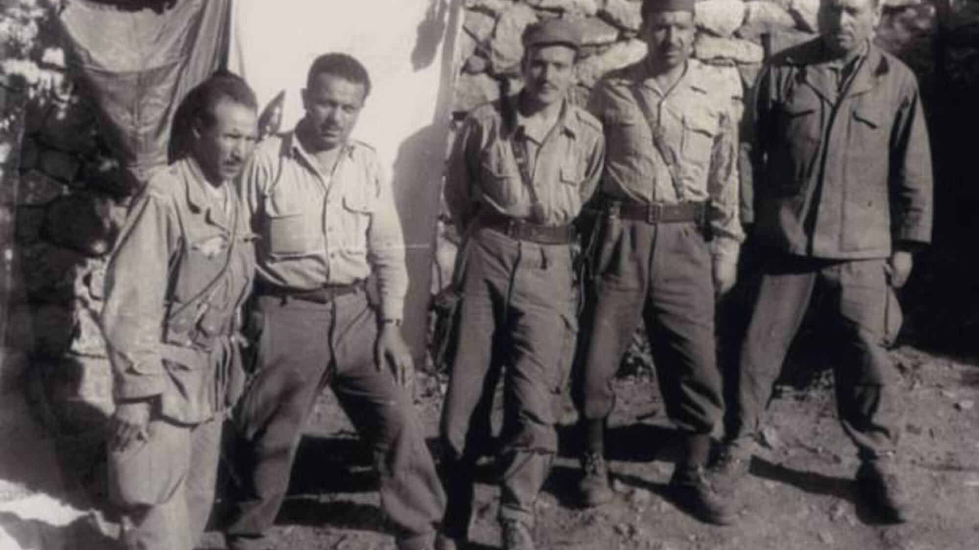 L'image présente un groupe de cinq hommes en uniforme militaire, debout ensemble devant un mur en pierres. Les hommes semblent être d'origine variée, portant des vêtements similaires, avec des pantalons et des chemises. Deux d'entre eux tiennent des armes à feu, et leur expression dégage un sentiment de camaraderie et de détermination. En arrière-plan, on aperçoit un drap suspendu, ce qui suggère qu'ils se trouvent dans un camp ou un endroit de rassemblement. L'atmosphère est sérieuse, typique des contextes militaires.