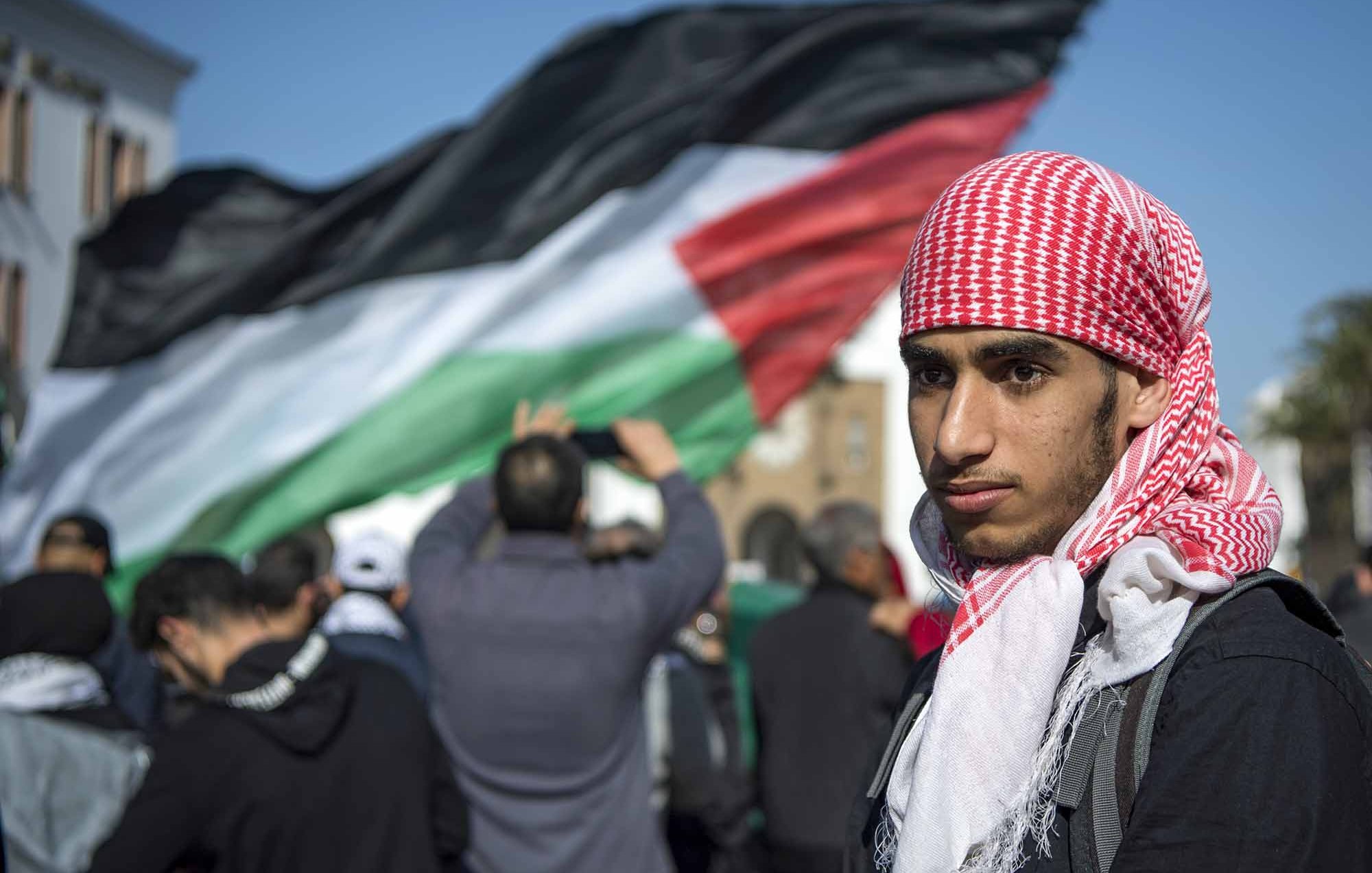 L'image montre un jeune homme portant un keffieh rouge et blanc, se tenant au premier plan. En arrière-plan, une grande foule est visible, avec un drapeau palestinien noir, blanc, vert et rouge qui flotte. L'atmosphère semble être celle d'une manifestation ou d'un rassemblement, où les participants expriment leurs convictions ou leur soutien à une cause. Le jeune homme affiche une expression sérieuse, ce qui ajoute à l'intensité de la scène.