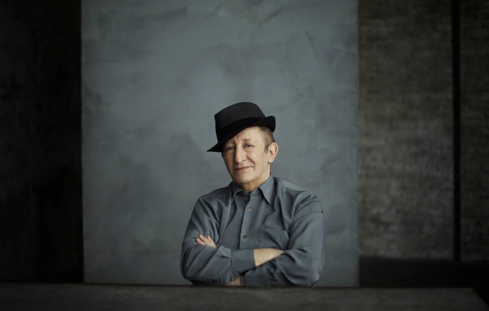 L'image montre un homme assis avec un chapeau noir. Il a un léger sourire et les bras croisés sur une table. Il porte une chemise grise. L'arrière-plan est simple, avec une texture de mur gris qui crée une atmosphère sobre et contemplative. La lumière est douce, accentuant les traits de son visage.