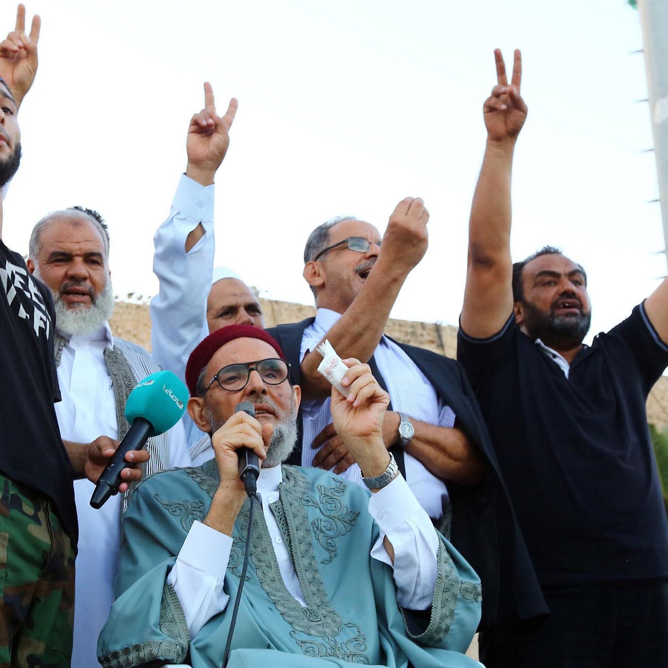 L'image montre un groupe d'hommes réunis lors d'un événement. Un homme au premier plan, portant une tenue traditionnelle, s'exprime tout en levant un symbole de paix avec ses mains. Les autres hommes en arrière-plan lèvent également les mains, probablement en signe de soutien ou de solidarité. L'ambiance semble festive et engagée, suggérant un moment de rassemblement ou de protestation. Des drapeaux ou des affiches pourraient être visibles, accentuant le contexte de l'événement.