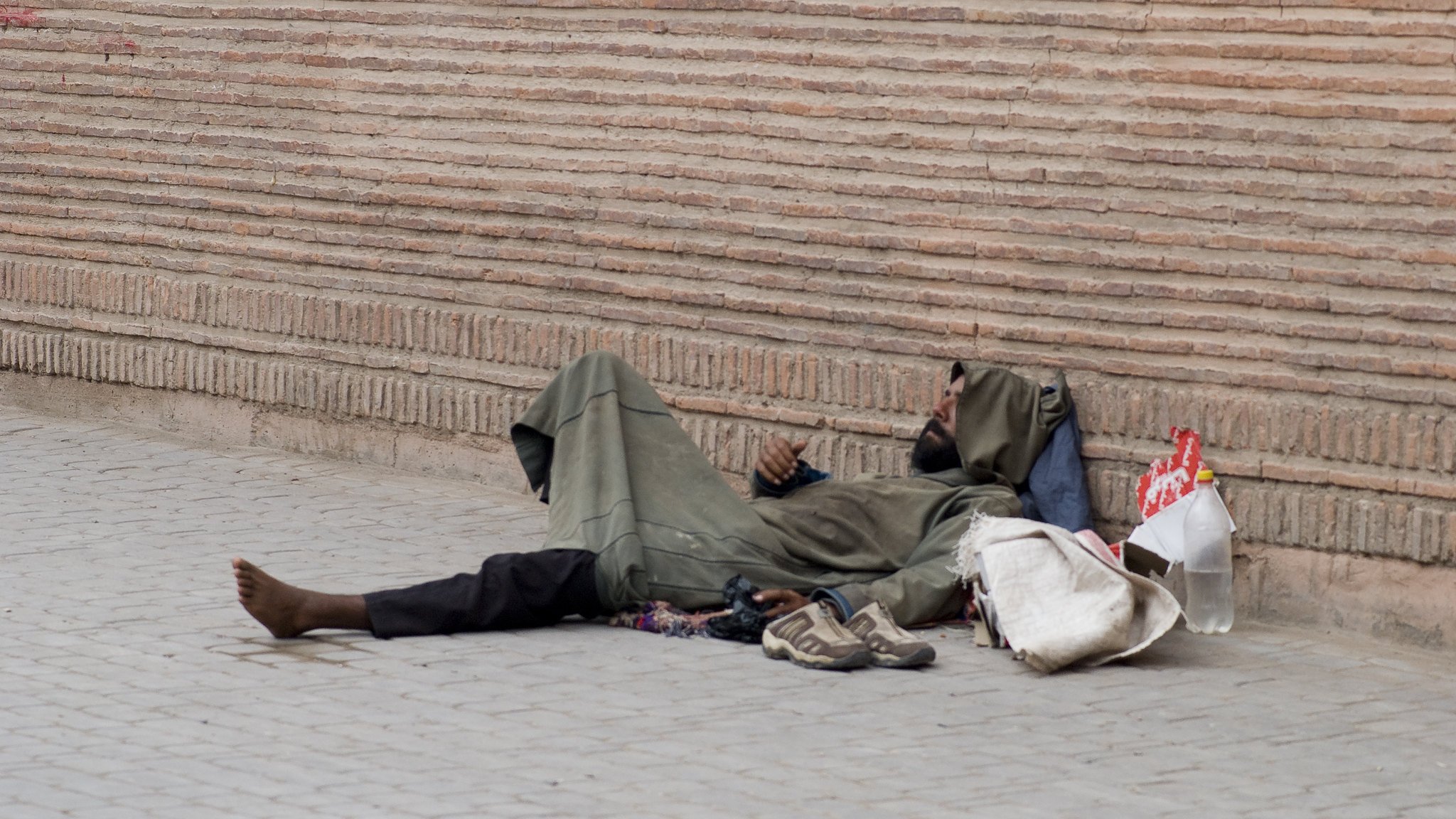 الصورة تظهر شخصًا ممددًا على الأرض، يرتدي ثيابًا بسيطة وفضفاضة. يبدو أنه في حالة استرخاء أو نوم. بجانبه بعض الأغراض مثل حذاء وحقيبة. الخلفية تتكون من جدار مصنوع من الطوب، مما يضيف إلى الأجواء الحضرية للصورة. المشهد يعكس حالة التحدي والمعاناة في الحياة اليومية لبعض الأفراد.