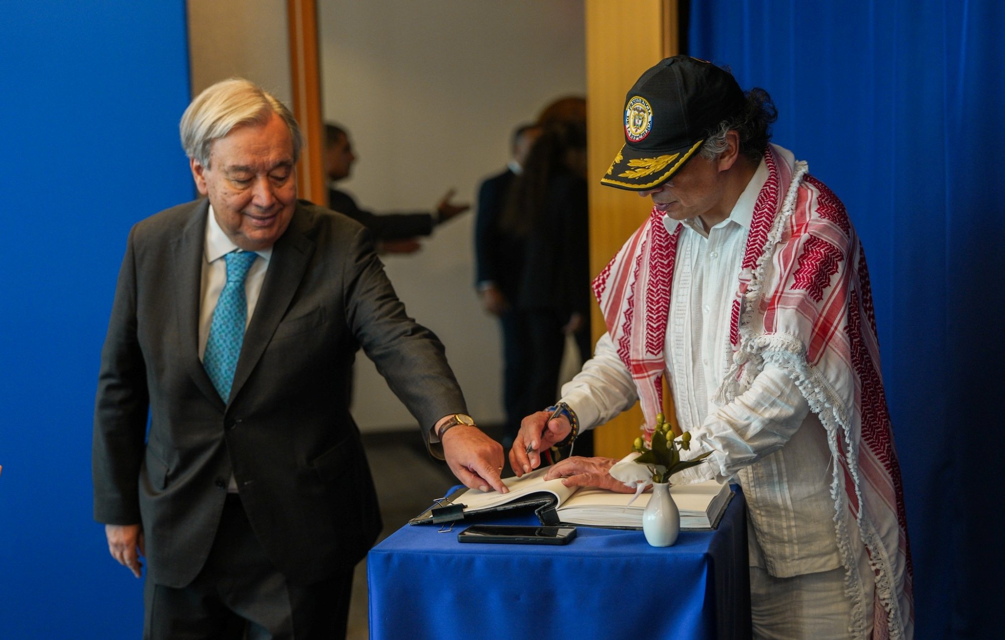 Two men are at a table; one is signing a book while the other observes. A blue backdrop is behind them.