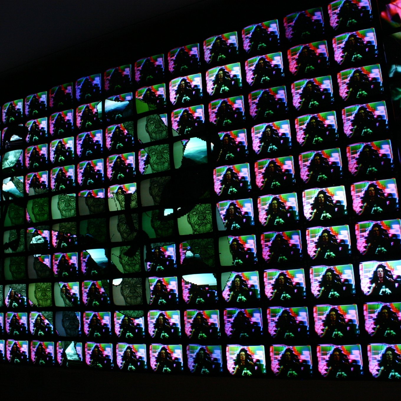 L'immagine mostra un ampio muro composto da molteplici schermi televisivi, ognuno dei quali visualizza immagini colorate e astratte. Le immagini si sovrappongono e si mescolano creando un effetto visivo vivace e dinamico. La combinazione di colori e forme sembra generare un'atmosfera futuristica e artistica, dando l'impressione di un'opera d'arte digitale contemporanea.