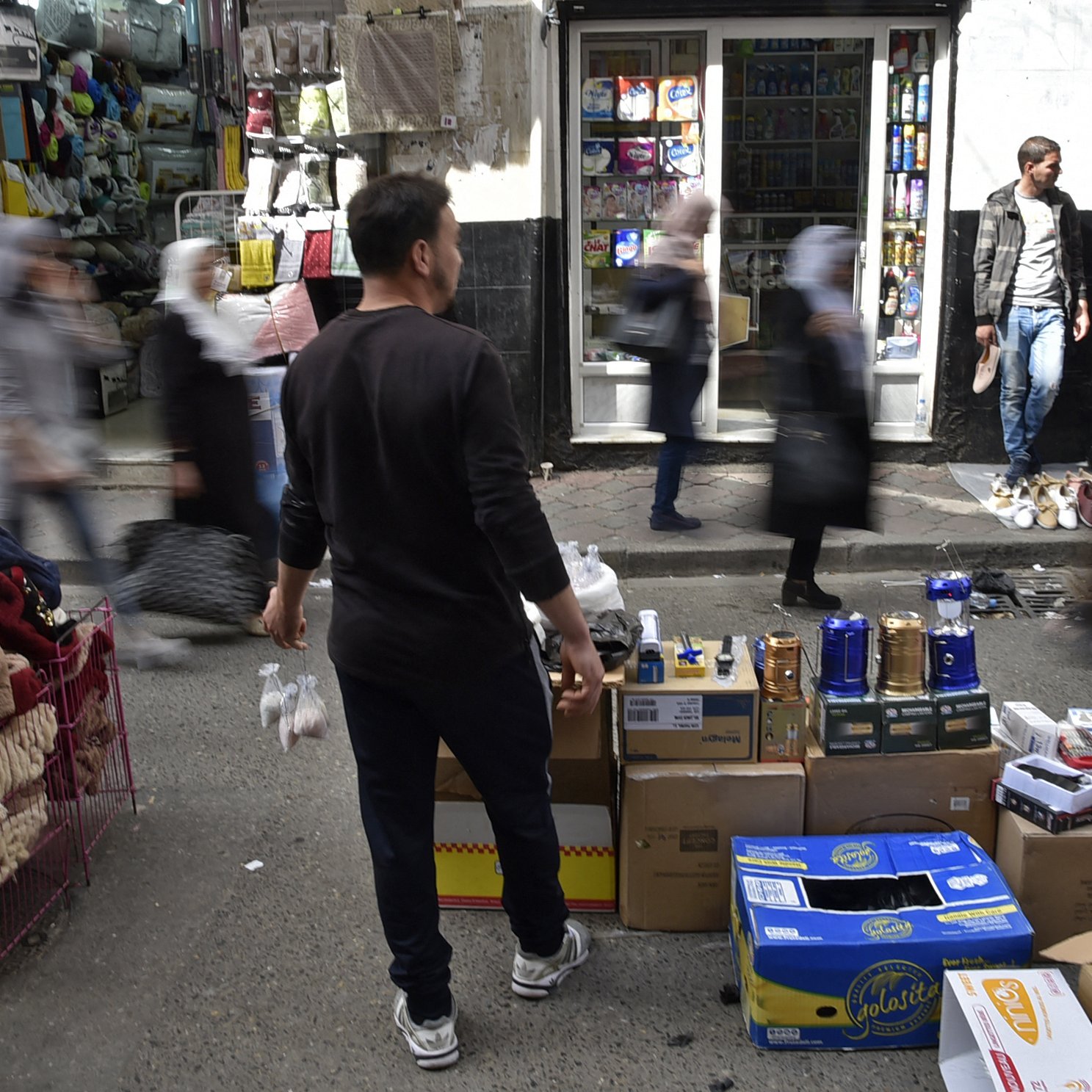 L'image montre une scène animée d'un marché urbain. On peut voir plusieurs personnes se déplacer rapidement, créant un effet de flou. Au premier plan, un homme se tient de dos, tenant des objets. Des étals sont visibles, avec des boîtes et des marchandises disposées sur des chariots. En arrière-plan, des magasins sont ouverts et on aperçoit des clients. L'atmosphère semble vivante et dynamique, typique d'un marché fréquenté.