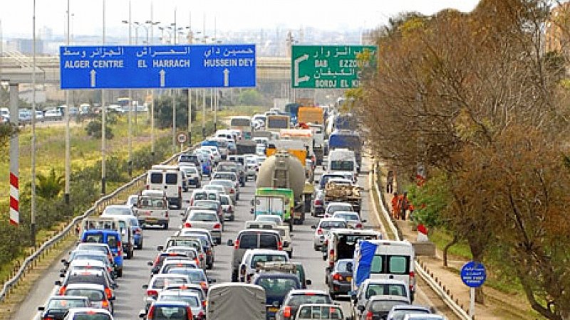 L'image montre une route très fréquentée avec une circulation dense. On peut voir de nombreux véhicules, y compris des voitures et des camionnettes, alignés les uns derrière les autres. Les panneaux de signalisation, écrits en arabe et en français, indiquent différentes destinations, comme "Alger Centre" et "Bab El Zawiya". Au loin, on aperçoit des arbres sur le bord de la route, ce qui suggère un environnement urbain. L'atmosphère semble être celle d'une journée ordinaire avec des bouchons routiers.