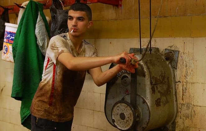 L'image montre un jeune homme dans un environnement de travail, probablement une boucherie ou un lieu de préparation alimentaire. Il est habillé de manière assez sales, avec des taches sur son t-shirt. Il tient un outil ou une manette pour actionner une machine, et il a une cigarette à la bouche. L'arrière-plan est constitué de murs carrelés avec des traces de saleté, créant une ambiance industrielle. L'atmosphère semble intense et le travail semble exigeant.