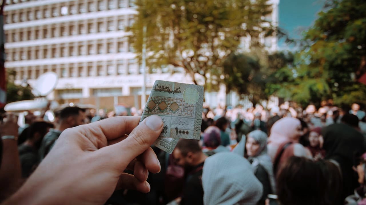 در این تصویر، یک دست در حال نگهداشتن یک کارت شناسایی یا مدرک مشابهی است. در پسزمینه، جمعیتی از افراد دیده میشود که در یک مکان عمومی جمع شدهاند. برخی از افراد چادر یا پوششهای اسلامی دارند. نور روز و فضای خارجی نشاندهنده یک رویداد یا تجمع عمومی است. احساس عمومی شلوغی و فعالیت اجتماعی در این تصویر مشهود است.