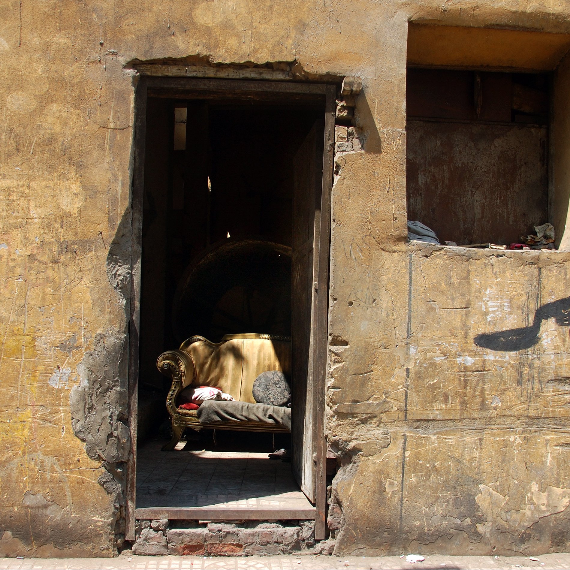 L'image montre une entrée partiellement ouverte d'un bâtiment avec un mur aux teintes jaunies et en mauvais état. À l'intérieur, on distingue un canapé doré, qui semble être un élément de mobilier abandonné. Le mur présente aussi des inscriptions en arabe, ajoutant une touche culturelle à la scène. L'ensemble dégage une atmosphère nostalgique, évoquant un espace autrefois habité et maintenant oublié.