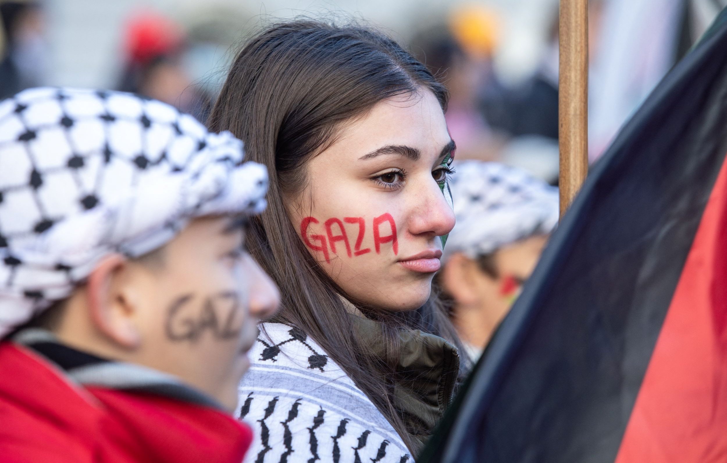 L'image montre des manifestants participant à une manifestation, probablement en lien avec la situation à Gaza. Une jeune femme a le mot "Gaza" peint en rouge sur sa joue, tandis qu'une autre personne à côté d'elle porte un foulard traditionnel palestinien et a également "Gaza" écrit sur son visage, mais en noir. Ils sont entourés d'un public qui semble mobilisé pour une cause. L'atmosphère est sérieuse, reflétant un engagement fort pour la situation en Palestine.