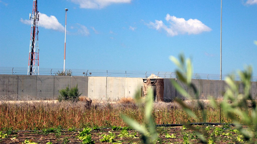 L'image montre un paysage avec un mur en béton qui semble délimiter une zone. Au fond, on peut voir des pylônes de communication et des lampadaires, suggérant une certaine infrastructure. Au premier plan, il y a un champ de cultures avec des lignes de légumes, probablement dans un état de sécheresse ou de négligence, tandis qu'un feuillage flou dans la partie inférieure de l'image ajoute une touche naturelle. Le ciel est bleu avec quelques nuages.