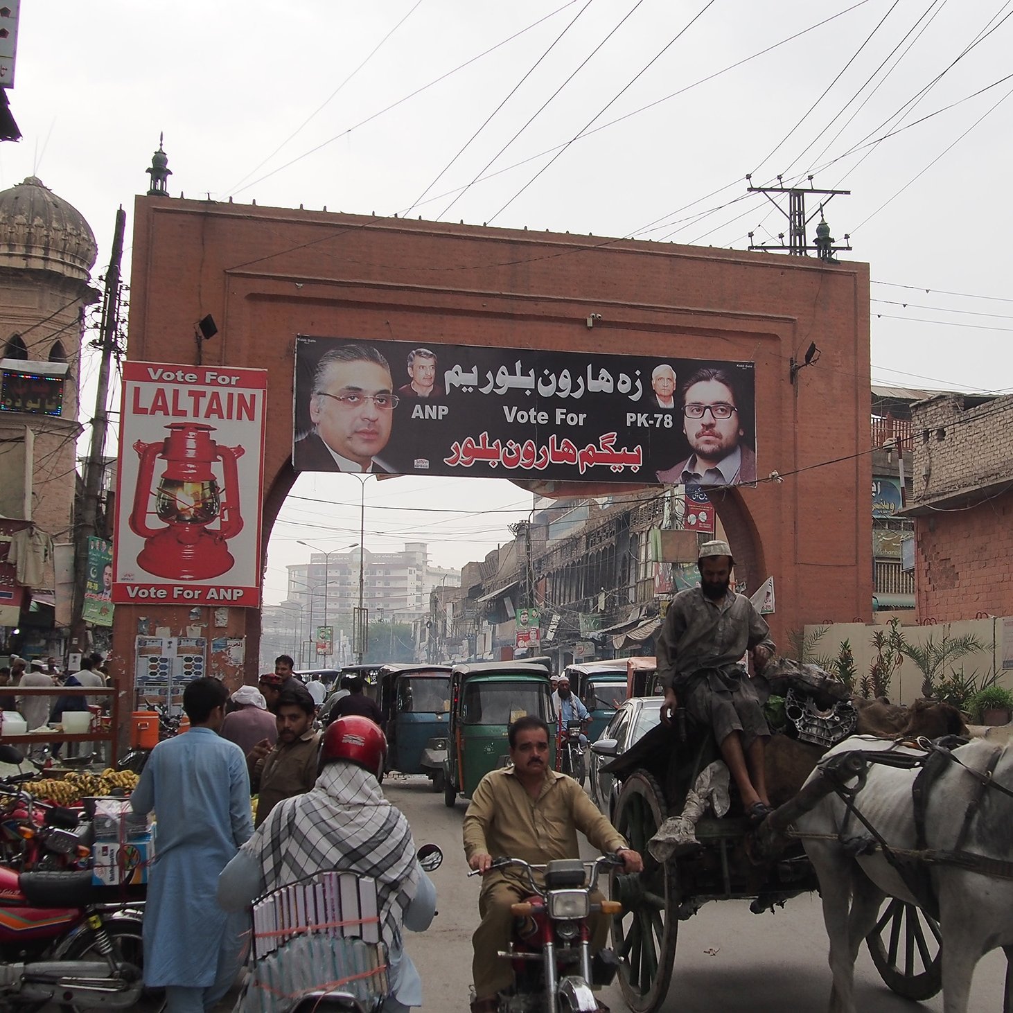 L'image montre une rue animée d'une ville avec un grand panneau publicitaire suspendu au-dessus. Ce panneau affiche des portraits de candidats politiques, accompagnés d'un message de campagne. Au premier plan, on voit des véhicules variés, y compris des charrettes tirées par des chevaux, des motos et des bicyclettes, illustrant l'activité de la vie urbaine. En arrière-plan, on peut apercevoir des bâtiments typiques et une ambiance dynamique, avec des personnes se déplaçant dans la rue.