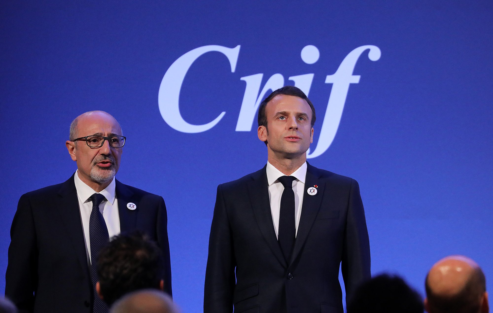 L'image montre deux personnes sur scène, probablement lors d'un événement officiel. À gauche, un homme porte des lunettes et un costume sombre, tandis qu'à droite, un homme, probablement un dirigeant, se tient debout en costume noir avec une cravate, affichant une expression sérieuse. En arrière-plan, le mot "Crif" est visible, ce qui suggère que l'événement est lié à cette organisation. L'atmosphère semble solennelle et professionnelle.