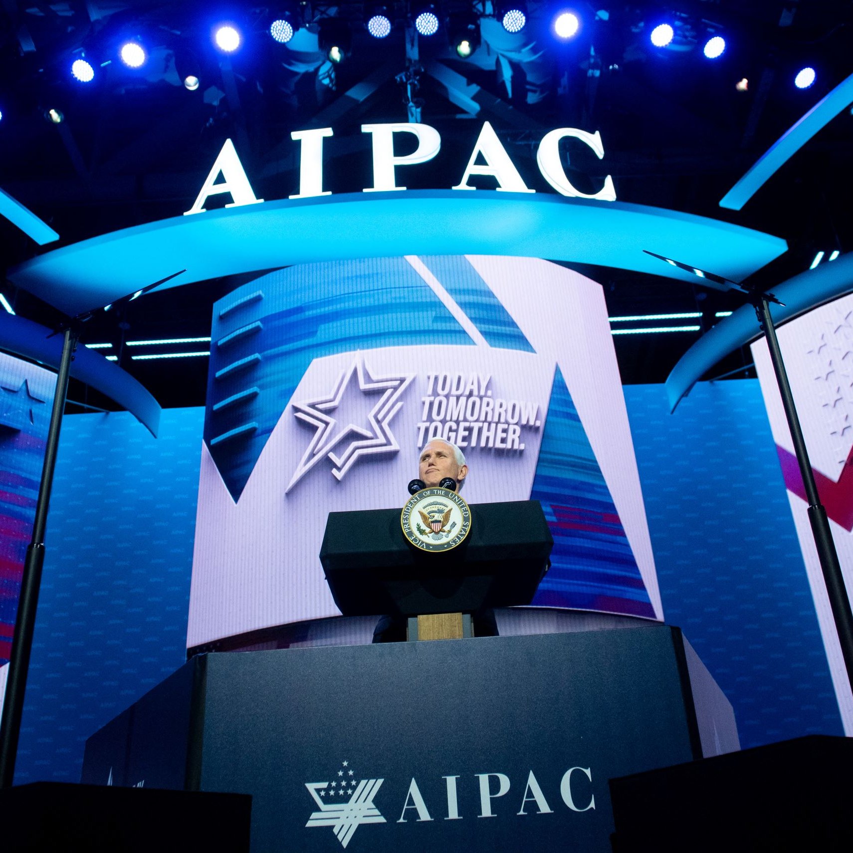 L'image montre une scène de conférence avec un podium bien en vue, sur lequel se trouve un sceau officiel. En arrière-plan, on peut voir un grand écran affichant le logo de l'AIPAC (American Israeli Public Affairs Committee), ainsi qu'un slogan qui pourrait être une thématique de l'événement. L'éclairage est dynamique, avec des lumières bleues qui créent une ambiance visuelle engageante. Cette configuration suggère qu'il s'agit d'un discours ou d'une présentation importante.