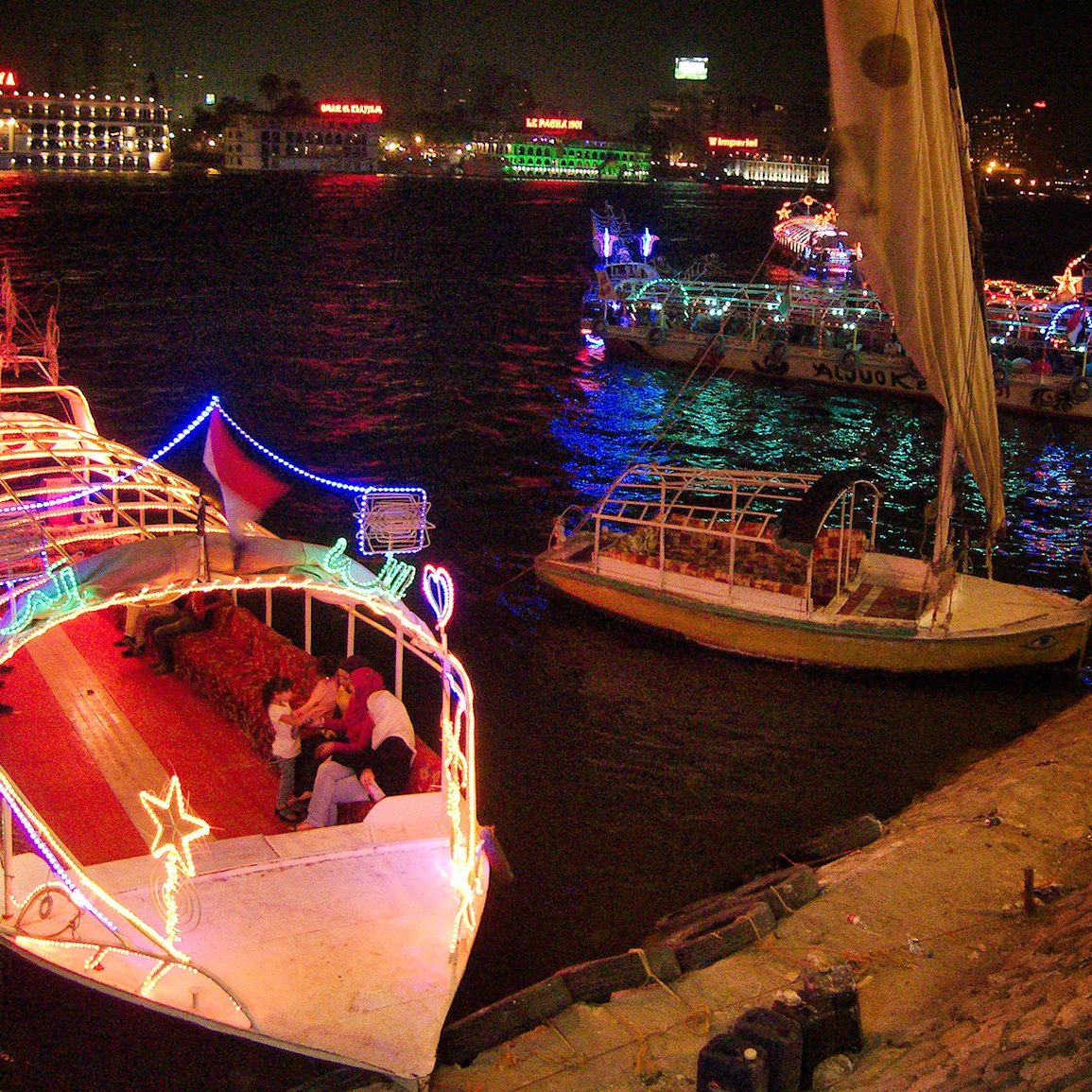 L'image montre un paysage nocturne animé le long d'une rivière. Plusieurs bateaux décorés de lumières colorées sont amarrés, créant une atmosphère festive. On peut voir des gens sur les bateaux et le long de la promenade, profitant de l'ambiance. En arrière-plan, des bâtiments illuminés ajoutent à la scène, avec des reflets dans l'eau. L'ensemble évoque une belle soirée en bord de rivière.