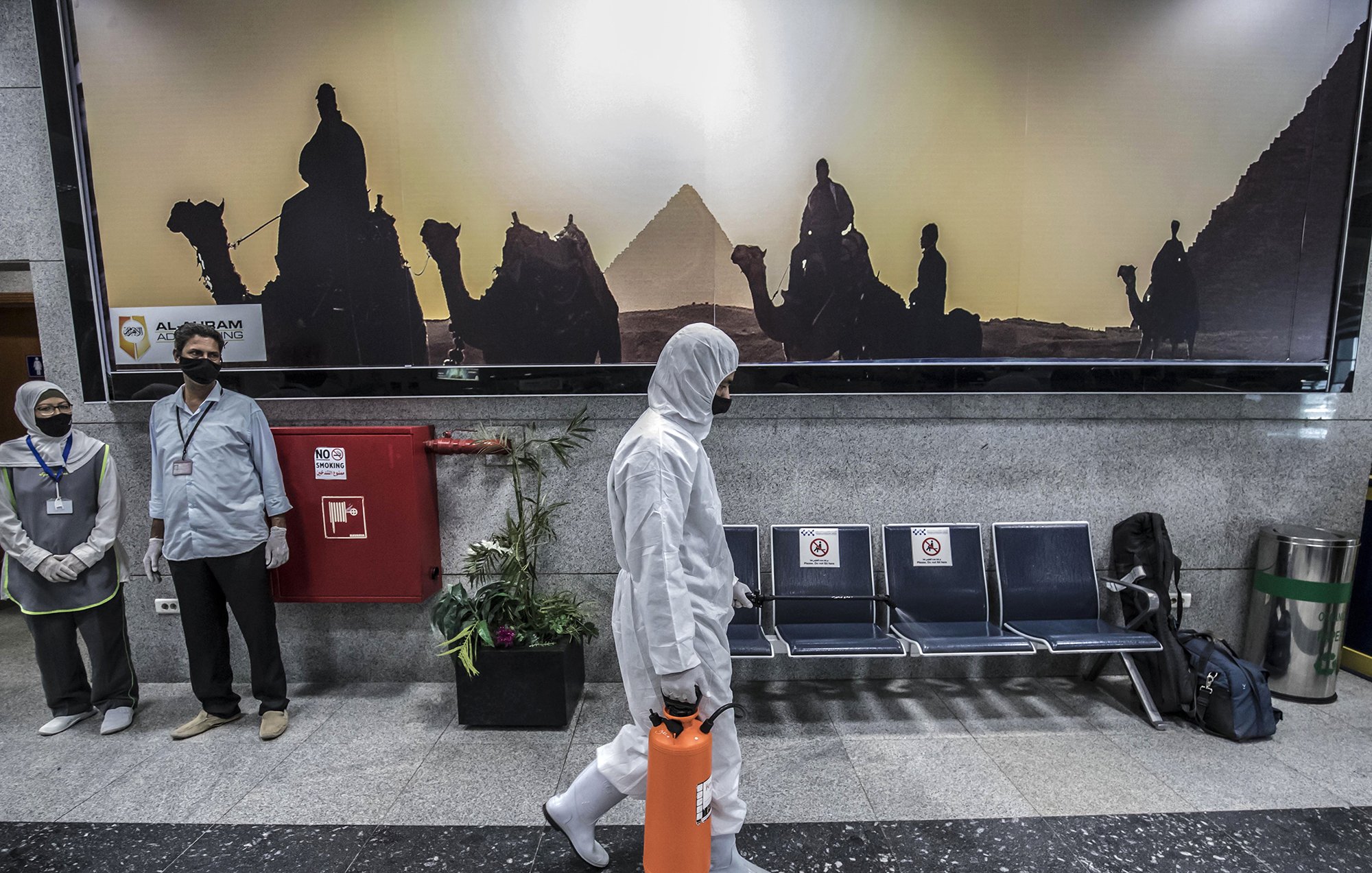 در این تصویر، یک فرد در حال قدم زدن است که لباسProtective پوشیده و یک جعبه نارنجی رنگ در دست دارد. در پس‌زمینه، تصویری از اهرام مصر و افرادی که بر روی شترها نشسته‌اند، به صورت سیاه و سفید دیده می‌شود. همچنین، دو نفر دیگر در سمت چپ تصویر ایستاده‌اند و ماسک زده‌اند. فضای کلی تصویر گویا محیطی عمومی یا فرودگاهی است.