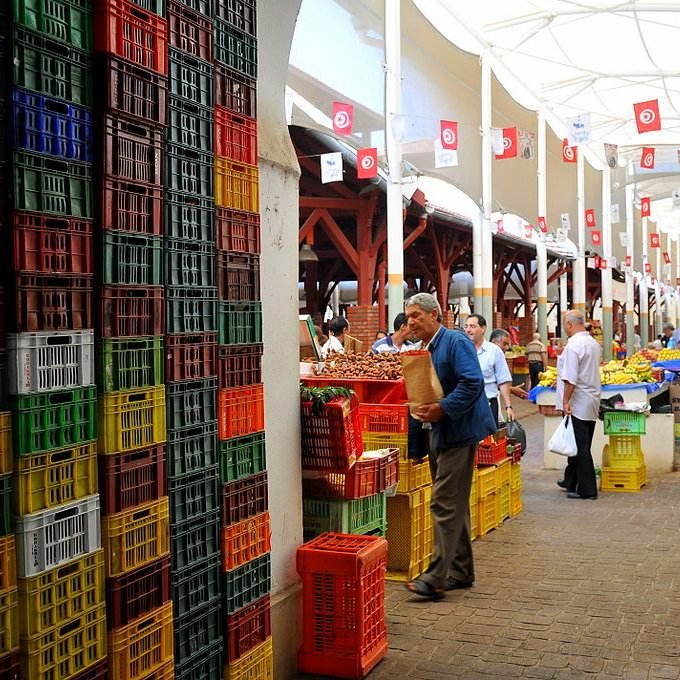 L'image montre un marché couvert animé, avec des étals bien organisés. On peut y voir des rangées de caisses colorées, de différentes tailles et en plastique, empilées de manière ordonnée. Des personnes se déplacent dans le marché, certaines portant des sacs, tandis que d'autres semblent inspecter les produits disponibles. Le tout est sous une structure avec un toit, permettant de se préserver du soleil. Des drapeaux tunisiens décorent le plafond, ajoutant une touche locale à l'ambiance du lieu.