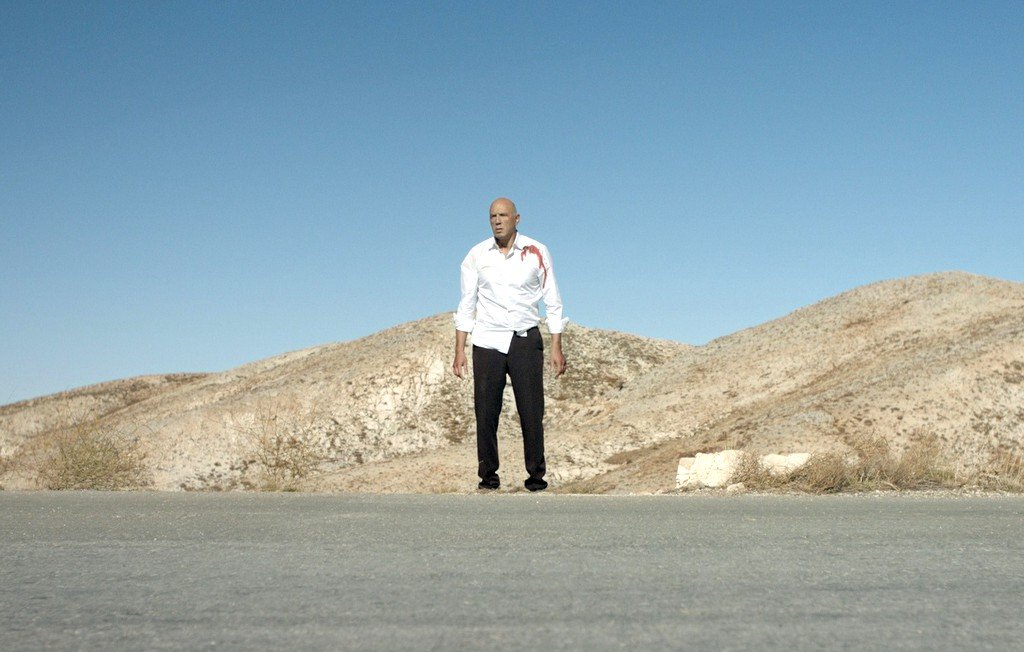 L'image montre un homme debout au milieu d'une route désertique, avec des collines arides en arrière-plan. Il porte une chemise blanche qui semble tâchée de sang et un pantalon noir. Le ciel est clair et bleu, ce qui souligne l'ambiance isolée et dramatique de la scène. Les éléments autour de lui donnent une impression de solitude et de tension.
