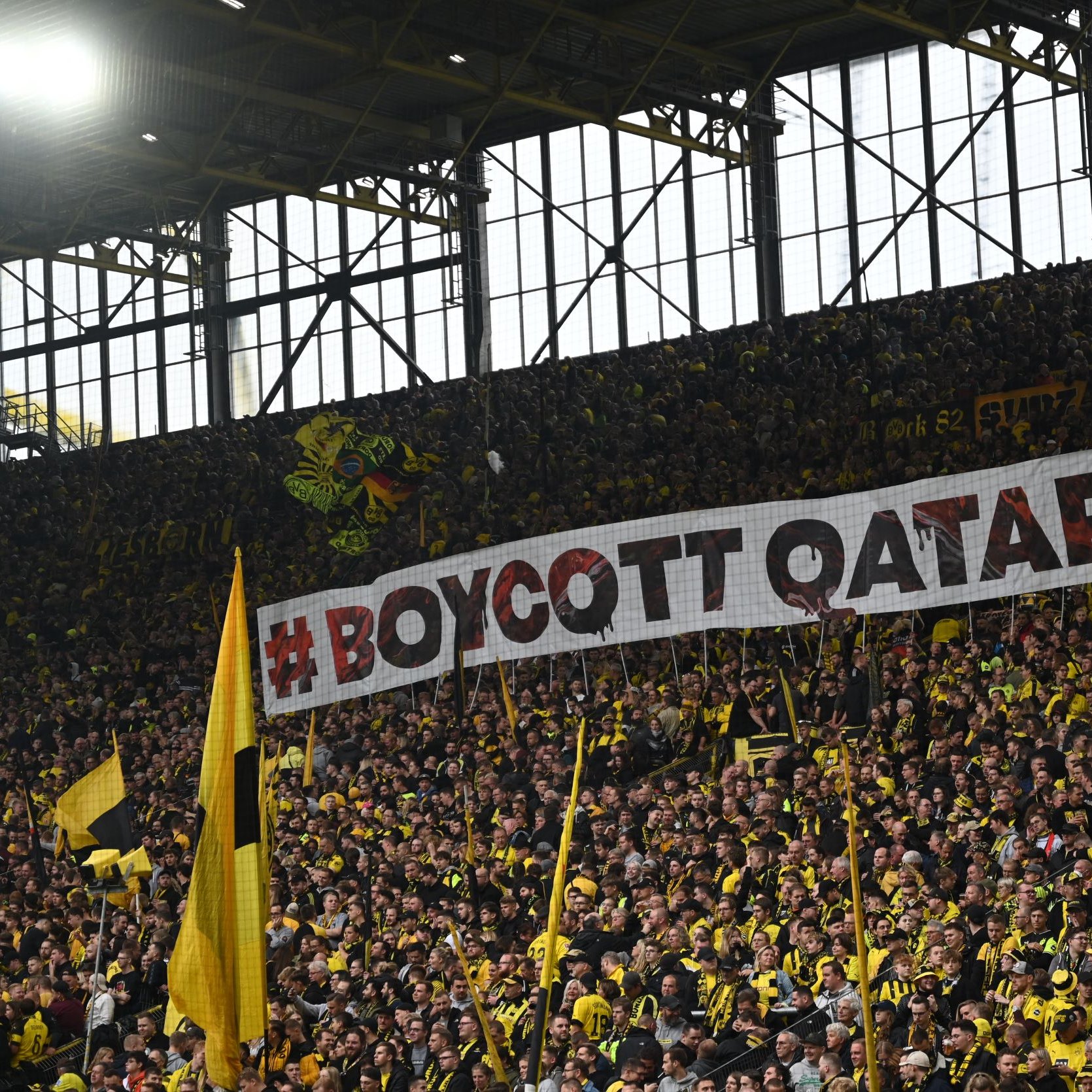 L'image montre un grand groupe de supporters dans un stade, portant des vêtements jaunes et noirs. Ils tiennent une immense banderole sur laquelle est inscrit "#BOYCOTT QATAR 2022". L'atmosphère semble dynamique et engagée, avec un fort sentiment de solidarité parmi les fans. Les tribunes sont pleines, créant une ambiance électrisante.