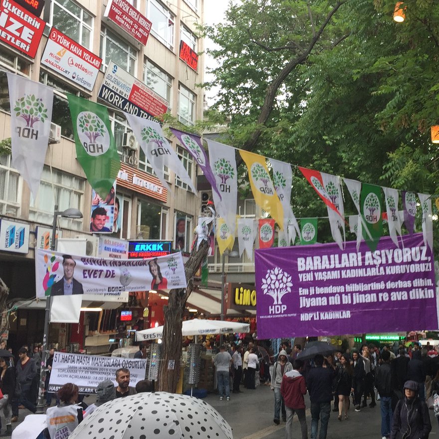 L'image montre une rue animée avec des banderoles accrochées entre les bâtiments. Ces banderoles semblent promouvoir un événement ou une cause, avec des slogans en caractères visibles. On peut également voir des groupes de personnes se rassembler au sol, suggérant une atmosphère de manifestation ou de réunion. Des arbres bordent la rue, ajoutant une touche de verdure à la scène. La journée semble nuageuse, ce qui pourrait indiquer un temps frais.