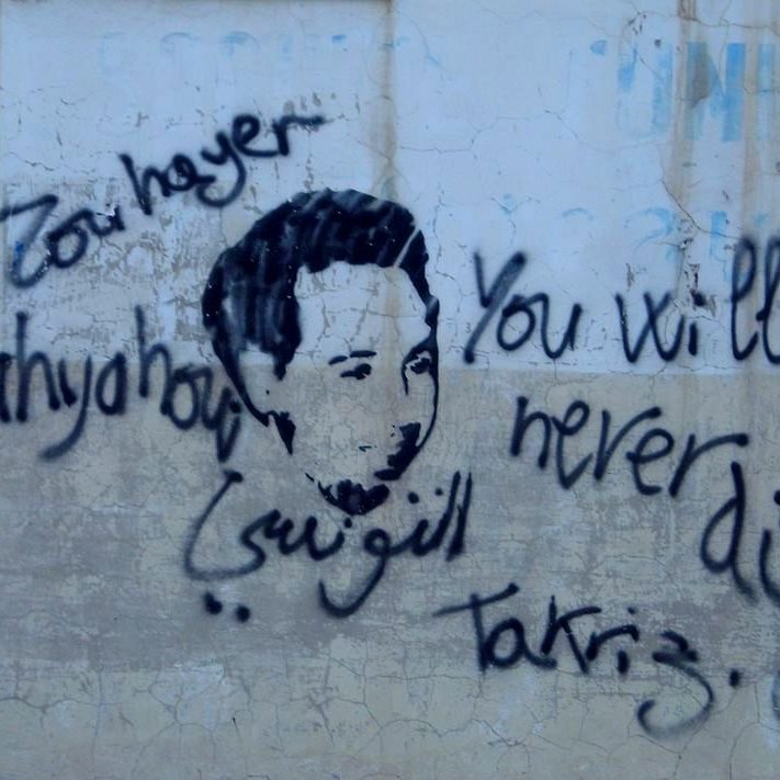 L'image représente un graffiti sur un mur. On y voit un visage stylisé, probablement d'une personne, entouré de mots écrits à la main. En haut, il y a le texte en arabe "Zou haya" et "Yahya", et en bas, le message en anglais "You will never die", accompagné du mot "Takríz" et du mot "Gun". L'ensemble dégage un sentiment de mémoire ou d'hommage, avec un mélange de langues.