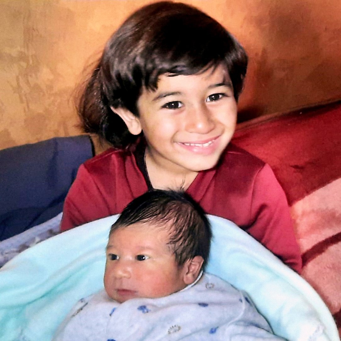 Nell'immagine si vede un bambino più grande che sorride mentre tiene in braccio un neonato avvolto in una coperta blu. Il bambino più grande ha i capelli scuri e lunghi, e indossa una maglietta rossa. Il neonato ha i capelli scuri e un'espressione curiosa. Sullo sfondo ci sono delle coperte sul lettino, creando un'atmosfera accogliente e familiare.