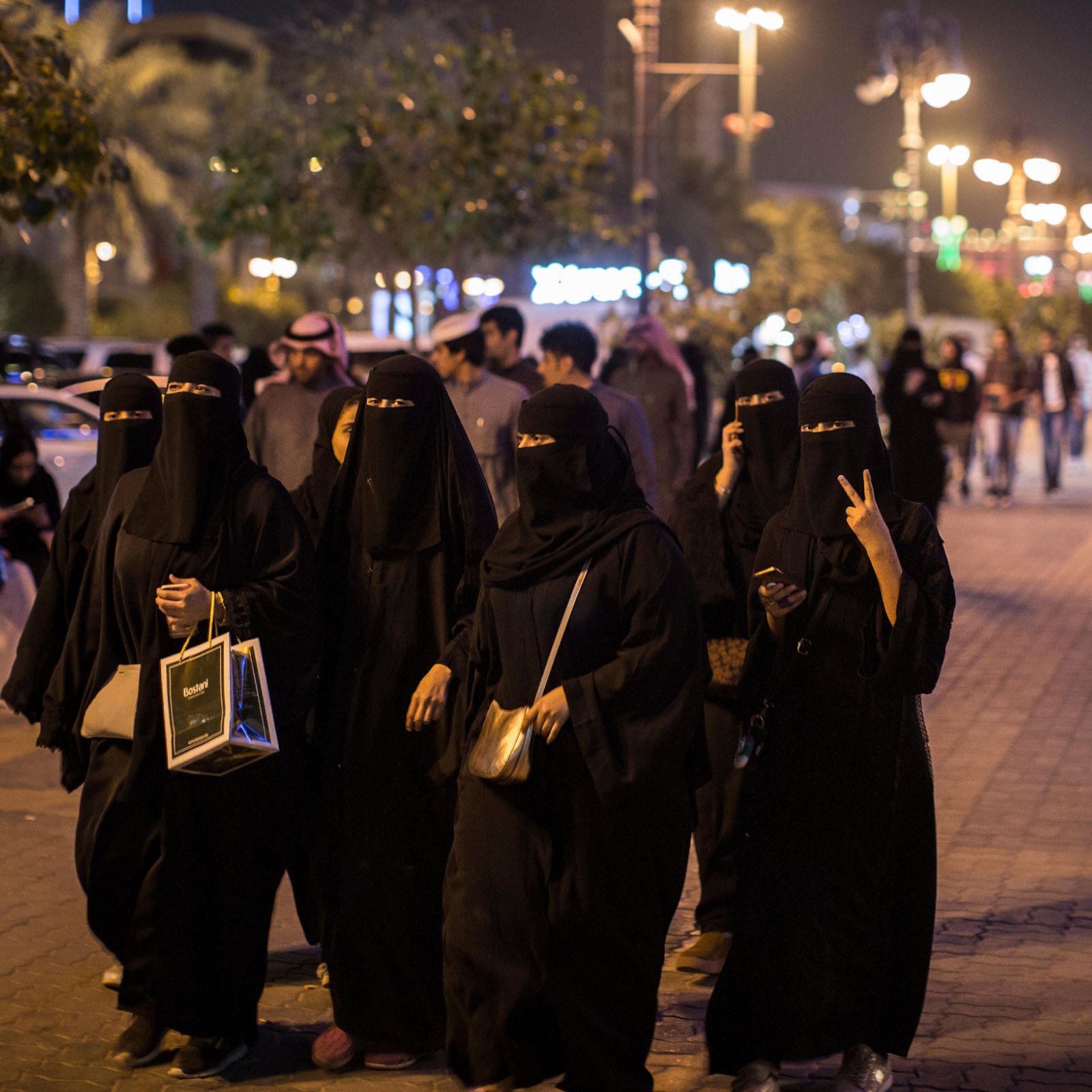 L'image montre un groupe de femmes vêtues de tenues noires traditionnelles marchant ensemble sur un trottoir. Certaines d'entre elles portent des masques qui dissimulent leur visage, tandis que d'autres affichent des gestes amicaux. En arrière-plan, on voit une rue animée avec des passants et des lumières, évoquant une ambiance nocturne. Un homme à vélo passe à proximité, ajoutant à la dynamique de la scène.