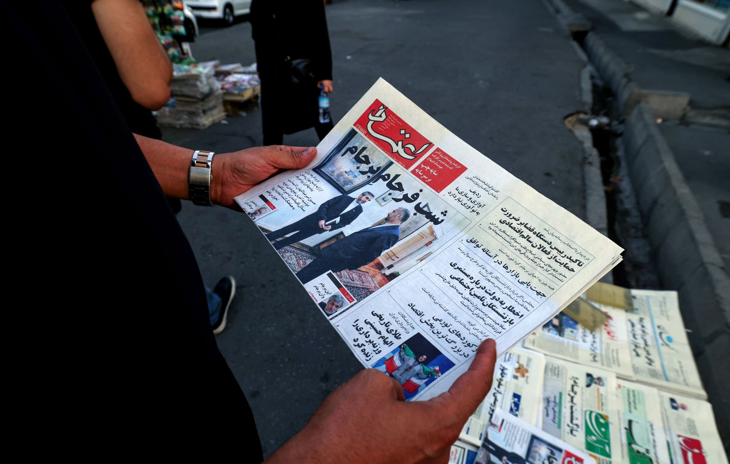 في الصورة، يُظهر شخص يحمل صحيفة باللغة العربية. تبدو الصحيفة جديدة ومطوية، ويظهر عنوان بارز على الصفحه الأولى، كما تُلاحظ مجموعة من الصحف في الخلفية. يبدو أن المشهد يدور في مكان عام، ربما في الشارع، حيث توجد بعض السيارات والأشخاص في محيط الصورة.