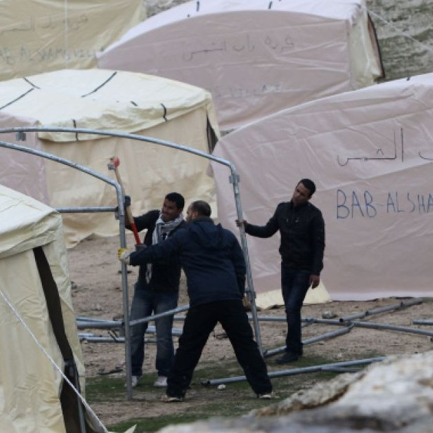 L'image montre plusieurs tentes de type campement, qui semblent être installées dans un environnement extérieur. Les tentes ont des écrits en arabe et en anglais, mentionnant "BAB AL SHAMS VILLAGE". On observe également des individus en train de travailler ensemble pour monter les structures des tentes. L'ensemble de la scène évoque des activités de construction ou d'aménagement dans un cadre probablement temporaire.