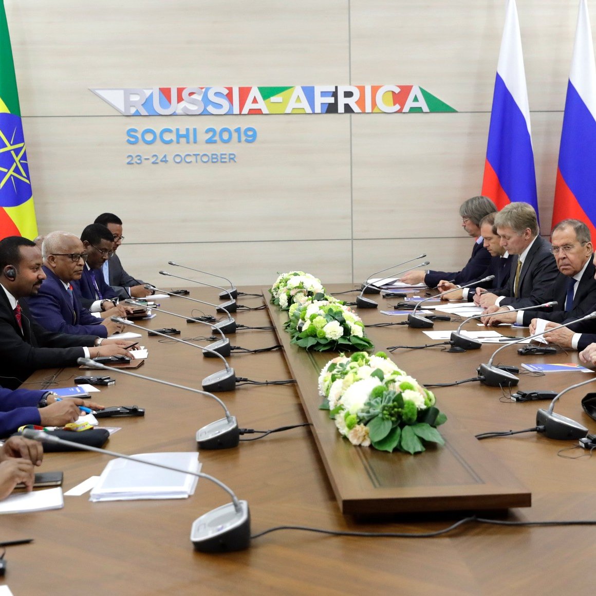 L'image montre une réunion officielle tenue à Sochi, en Russie, en octobre 2019, dans le cadre d'un sommet Russie-Afrique. On peut voir des représentants de plusieurs pays africains, ainsi que des dignitaires russes, assis autour d'une grande table de conférence. Des drapeaux représentant l'Éthiopie et un autre pays africain sont visibles en arrière-plan. Au centre de la table, il y a des arrangements floraux. Les participants semblent engagés dans une discussion, avec certains utilisant des écouteurs pour la traduction.