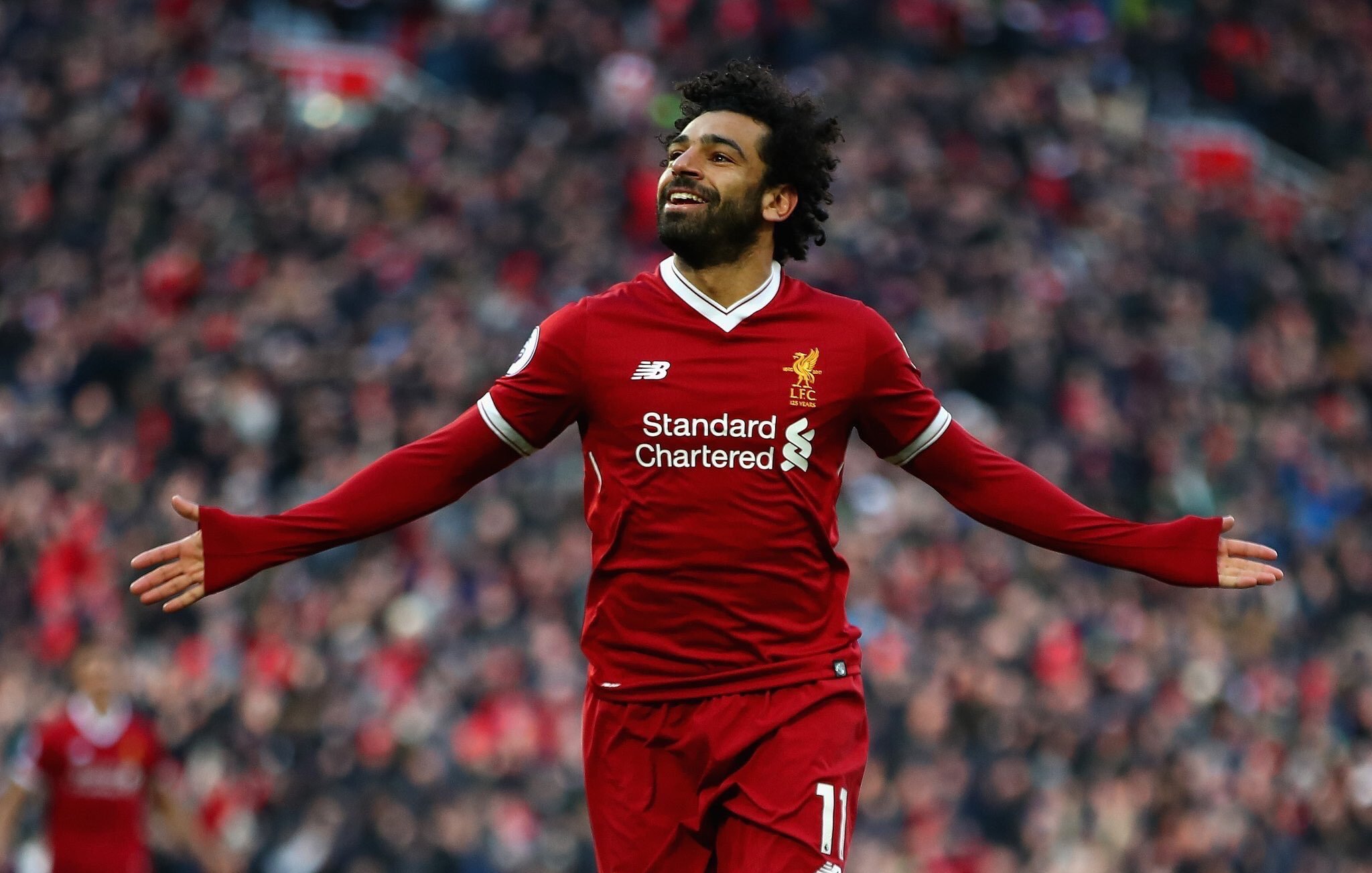 L'image montre un footballeur célébrant un but. Il porte un maillot rouge avec le logo de Liverpool FC, ainsi que des logos de sponsors. Son expression est joyeuse, avec un grand sourire, et il étend les bras, manifestant sa joie devant les supporters. L'arrière-plan est flou, mais on peut deviner une foule enthousiaste dans un stade.