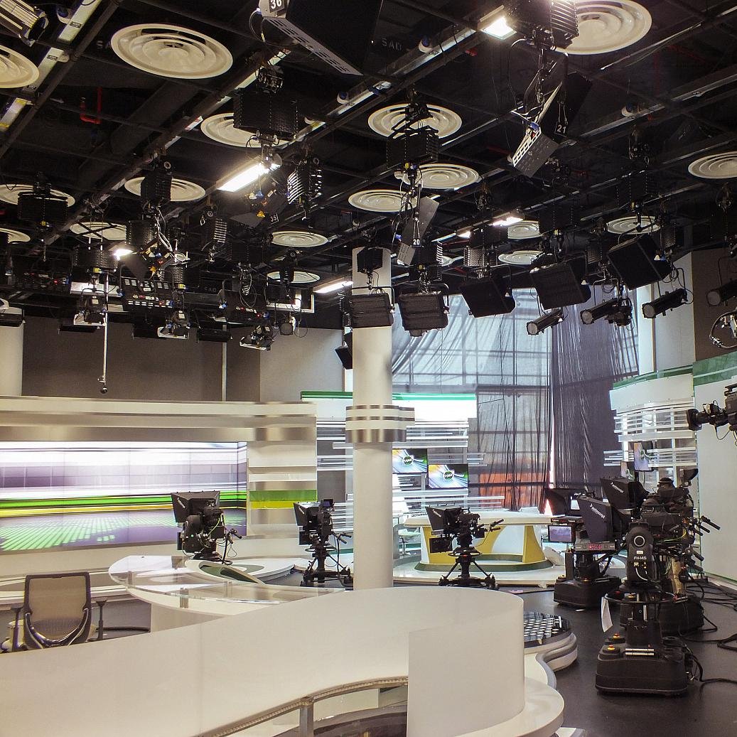 L'image montre un studio de télévision. On peut voir plusieurs caméras montées sur des tréteaux, prêtes à capturer des scènes. Le décor est moderne, avec des surfaces blanches et des accents de couleurs vives, notamment du vert. Des lumières suspendues au plafond éclairent le plateau, et il y a des écrans en arrière-plan, probablement utilisés pour afficher des informations ou des graphiques. L'ambiance semble professionnelle et organisée, typique d'un environnement de diffusion.