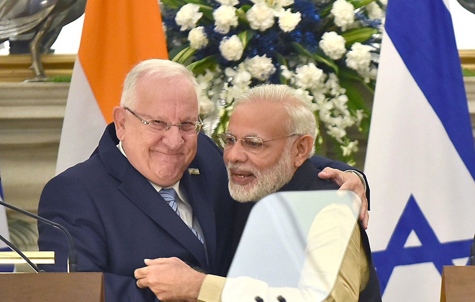L'image montre deux hommes en train de se serrer dans les bras, exprimant une atmosphère chaleureuse et amicale. L'un des hommes porte un costume sombre avec une cravate, tandis que l'autre est vêtu d'une tenue traditionnelle indienne. En arrière-plan, on peut voir des drapeaux de l'Inde et d'Israël ainsi que des fleurs décoratives, ce qui indique un contexte officiel ou diplomatique.