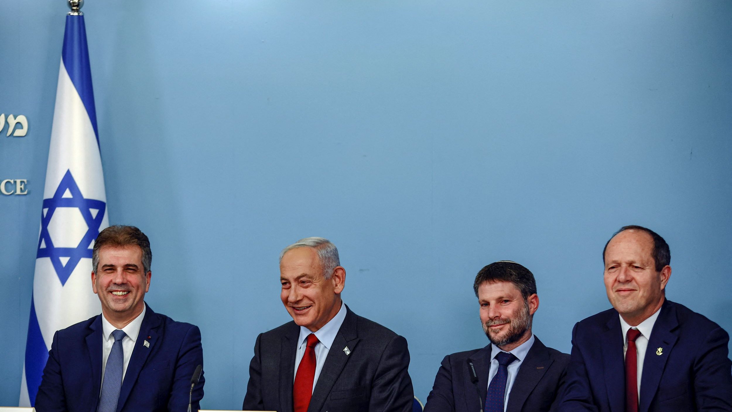 La imagen muestra a un grupo de cuatro personas sentadas alrededor de una mesa en un entorno oficial. Al fondo, se puede ver una bandera de Israel. Las personas parecen estar en un evento o una conferencia, y todos tienen expresiones serias pero amigables. Están vestidos de manera formal. La escena sugiere un contexto político o gubernamental.