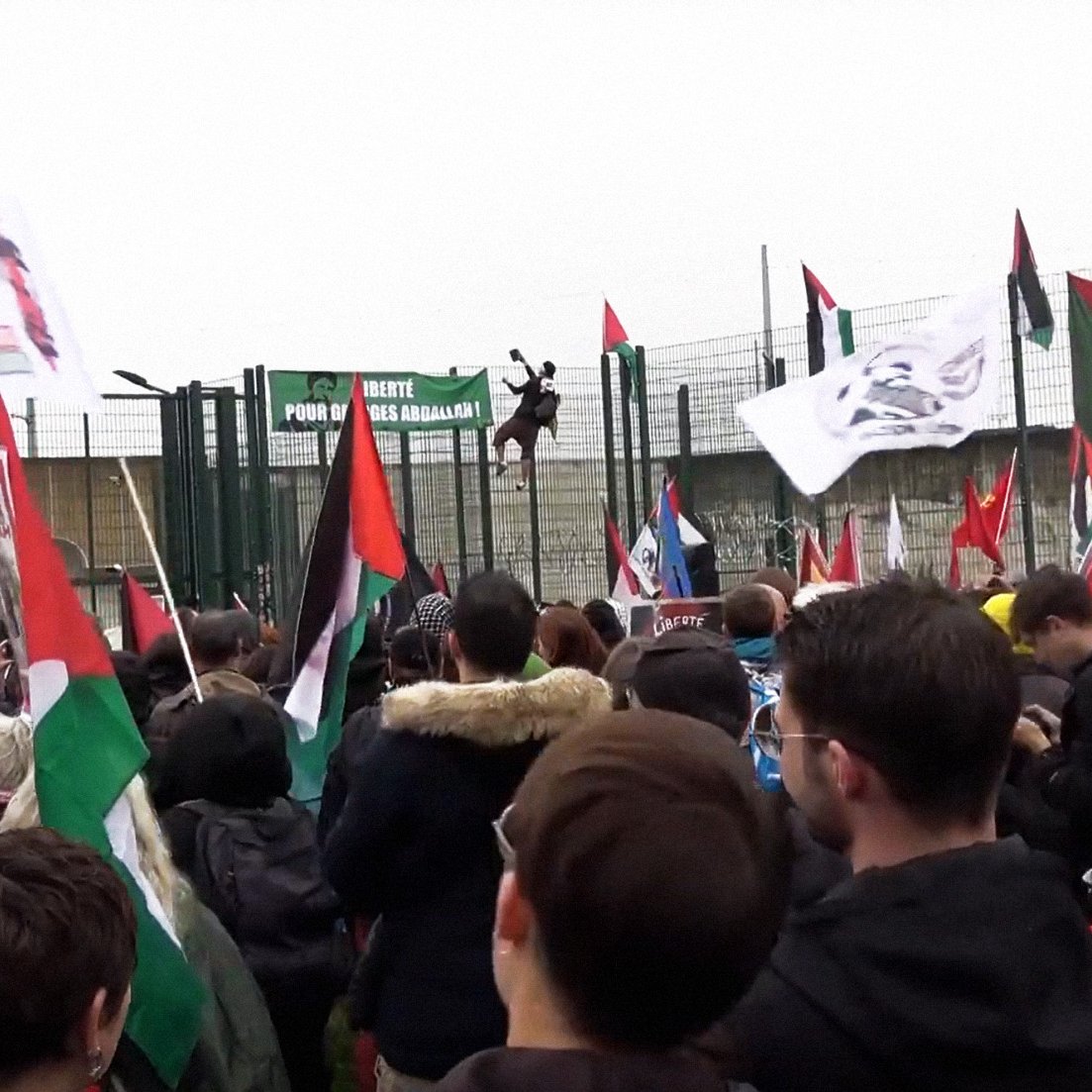 L'image montre une foule rassemblée lors d'une manifestation. Des personnes tiennent des drapeaux de différentes couleurs, dont des drapeaux palestiniens et d'autres symboles politiques. On peut voir une grande structure en arrière-plan, probablement une barrière ou une clôture, avec des bannières accrochées. L'atmosphère semble dynamique et engagée, reflétant une ambiance de protestation ou de revendication. Le temps paraît nuageux, ajoutant à l'intensité de l'événement.