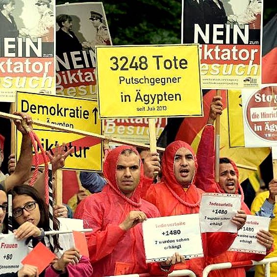 L'image montre un groupe de manifestants rassemblés pour une protestation. Ils portent des vêtements de pluie rouges et tiennent des pancartes avec des messages en allemand et en anglais, dénonçant une dictature et parlant de décès d'opposants en Égypte. On peut voir des slogans comme "NEIN" et des chiffres indiquant le nombre de morts. L'atmosphère semble sérieuse, témoignant de l'engagement des manifestants pour leurs causes.
