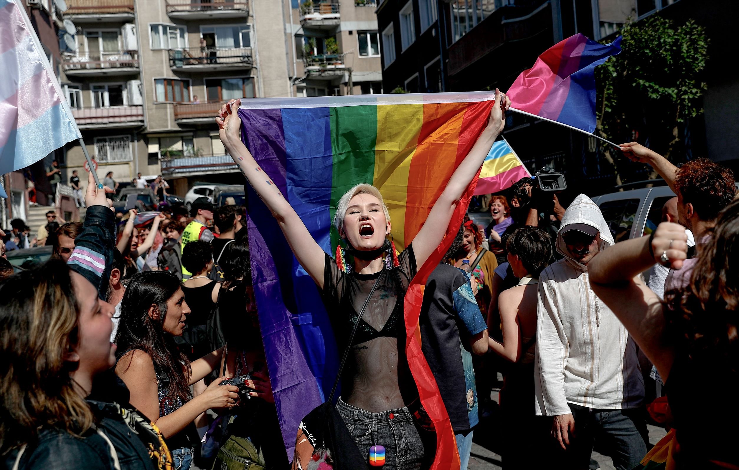 الصورة تظهر مجموعة من الأشخاص في تظاهرة للاحتفال بالفخر والمساواة. في المقدمة، هناك شخص يحمل علم قوس قزح، وهو رمز لحركة حقوق LGBTQ+. يبدو الشخص متحمسًا ويرتدي ملابس مميزة. الخلفية تحتوي على حشود من الناس وبعض الأعلام والمباني، مما يعكس أجواء الاحتفال والتعبير عن الهوية. التجمع يبدو مفعمًا بالحيوية والطاقة.