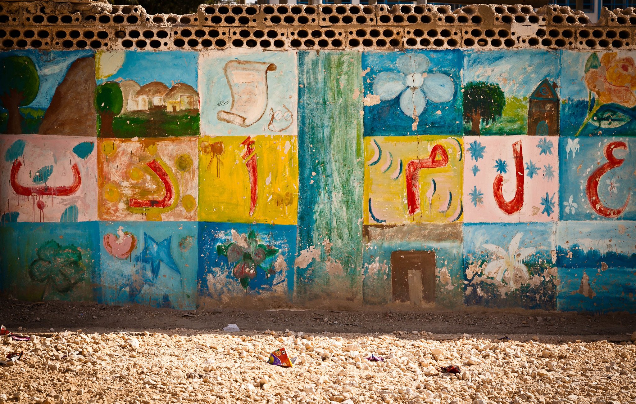 L'image montre un mur décoré de peintures colorées. Les motifs incluent des formes géométriques, des fleurs et des éléments abstraits. Les couleurs vives comme le bleu, le vert, le jaune et le rouge créent un contraste attrayant. Le bas du mur semble légèrement abîmé par le temps, et le sol est recouvert de petits débris, ce qui donne un aspect un peu désordonné à l'ensemble. Cette scène évoque un environnement créatif, peut-être dans un espace communautaire ou scolaire.