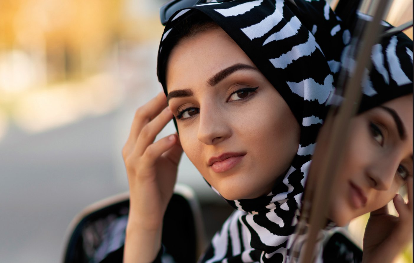 L'image montre une femme portant un hijab à motifs zebrés noirs et blancs. Elle se tient près d'une voiture, regardant vers la caméra avec une expression calme et confiante. Ses lunettes de soleil sont posées sur le haut de sa tête, ajoutant une touche de style. En arrière-plan, on peut apercevoir des contours flous d'un environnement urbain, suggérant un jour ensoleillé.
