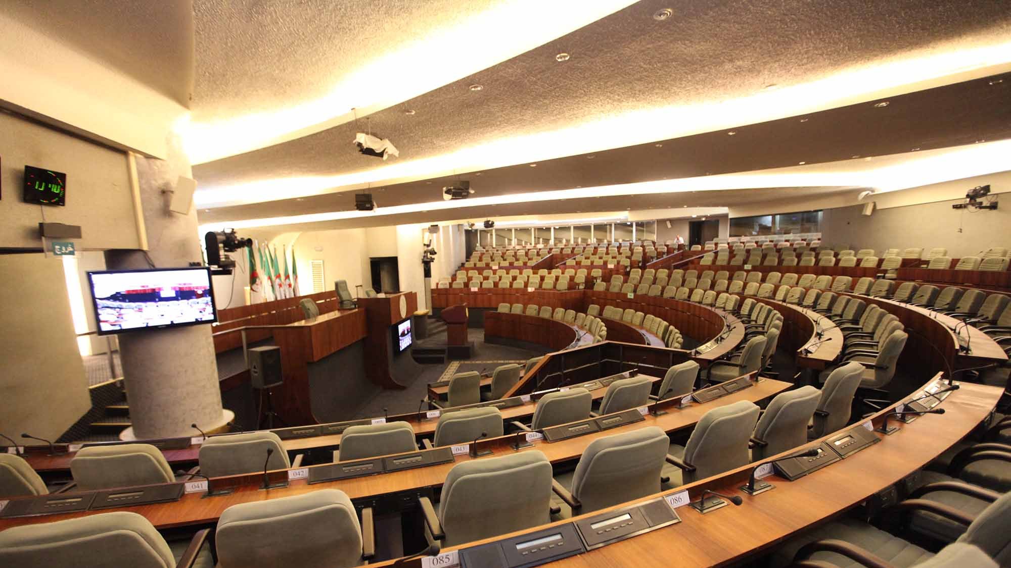 L'image représente une salle de réunion ou un hémicycle, typique des assemblées législatives. On peut voir des sièges disposés en amphithéâtre, avec des rangées de fauteuils gris. À l'avant, il y a une estrade avec un bureau et des équipements audiovisuels. La décoration est moderne et sobre, montrant un environnement formel adapté aux discussions et aux débats. Des écrans sont également présents pour fournir des informations aux membres présents.