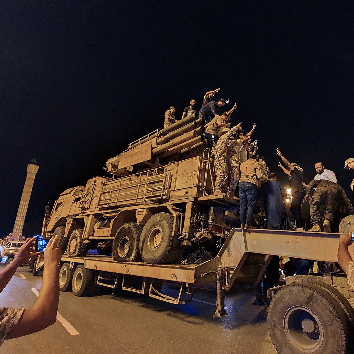 L'image montre un groupe de personnes rassemblées autour d'un véhicule militaire sur une route, probablement pendant la nuit. Plusieurs individus sont montés sur le véhicule et semblent exprimer des émotions fortes, peut-être en célébrant ou en manifestant un soutien. D'autres personnes prennent des photos avec leurs téléphones. L'ambiance générale semble dynamique et impliquée, avec une forte présence militaire sur le site.