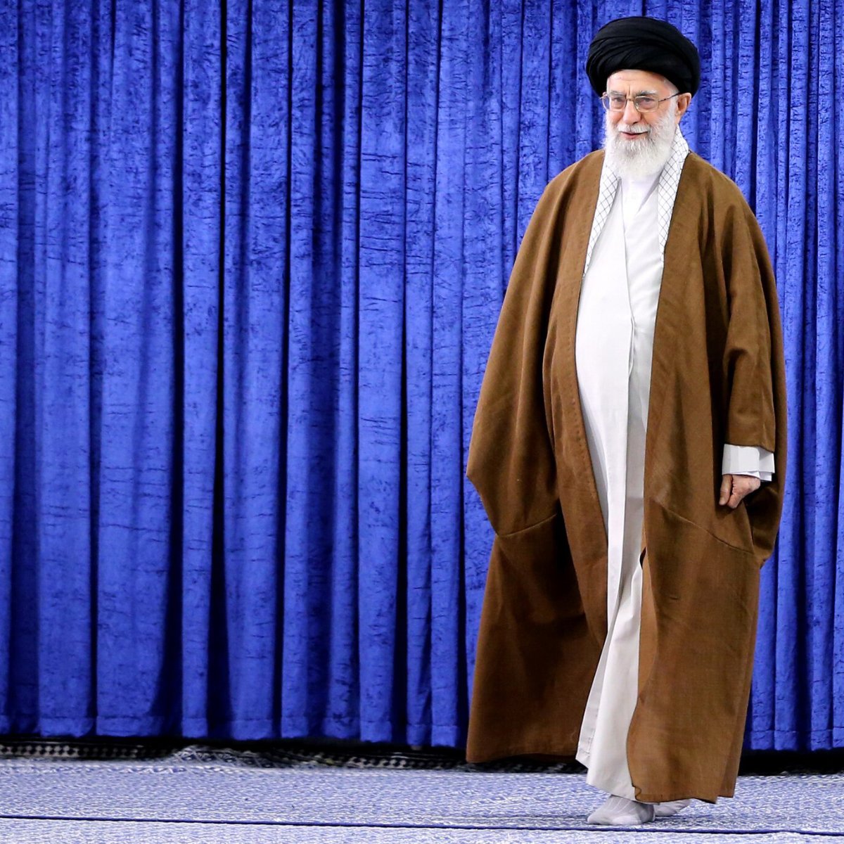 L'image montre une personne qui marche sur un tapis, devant un fond de rideaux bleus. Cette personne porte une longue robe traditionnelle brune et un turban noir. L'environnement semble être un espace formel, peut-être un studio ou une salle de réunion. Le ton de l'image est serein et solennel, mettant en avant la posture et l'attitude de la personne.