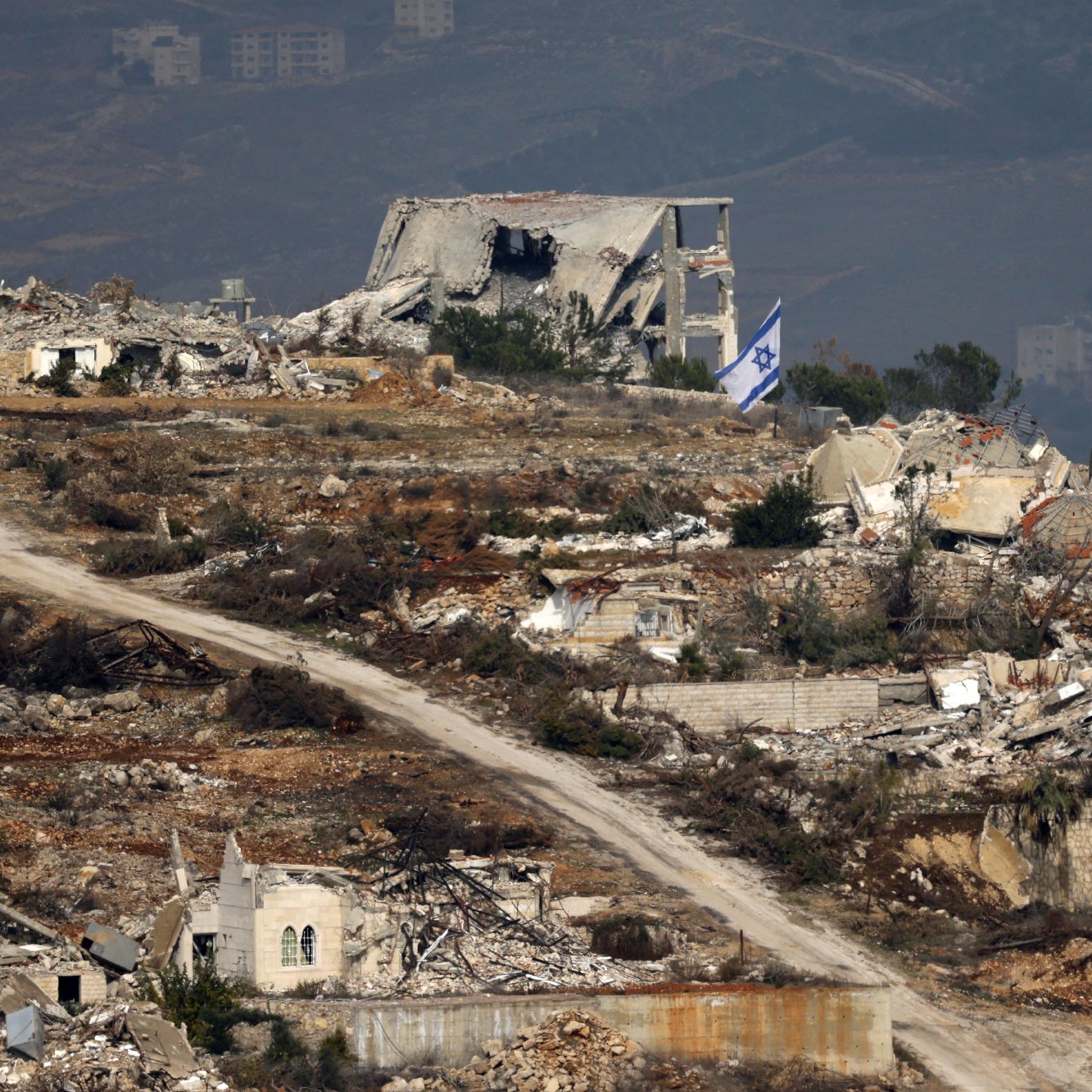 L'image montre un paysage de ruines, probablement dans une région touchée par un conflit. À l'arrière-plan, on peut voir des bâtiments détruits, avec des débris éparpillés. Une route de terre serpente à travers le paysage, et un drapeau israélien apparaît au premier plan, signalant la présence israélienne dans la zone. Le paysage est montagneux, avec des collines en arrière-plan, suggérant un environnement rural. L'image évoque une atmosphère de désolation et de conflit.